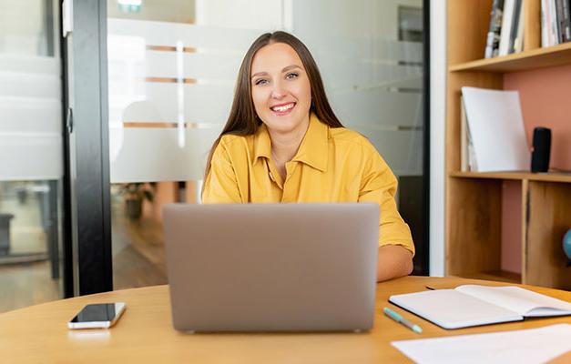 办公室里，穿着黄色衬衫的微笑女人坐在笔记本电脑后面